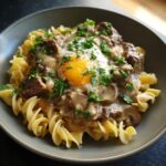 A bowl of Classic Beef Stroganoff with egg noodles, topped with a fried egg and parsley.