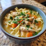 Close-up of a bowl of creamy chicken noodle soup with chicken, carrots, noodles, and parsley.