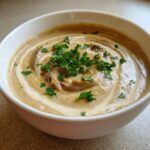 Close-up of a bowl of Creamy Mushroom Soup, swirled with cream and garnished with parsley.