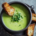 Close-up of a bowl of Creamy Pea & Mint Soup garnished with fresh herbs and served with toasted bread.
