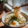 Close-up of a bowl of Garlic Miso Ramen with noodles being lifted by chopsticks, garnished with egg and scallions.