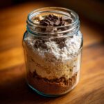 A clear glass jar filled with layers of cookie mix ingredients, topped with chocolate chunks. This is a giftable cookie mix in a jar.