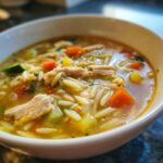Close-up of a bowl of delicious Lemon Chicken Orzo Soup, with chicken, orzo, and vegetables.