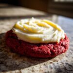 A close-up of a vibrant red velvet cookie topped with swirls of creamy white frosting.