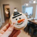A hand holding an adorable Snowman Hat Cookie decorated with white icing, black hat, orange carrot nose, and black eyes.