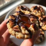 Close-up of a hand holding a Turtle Thumbprint Cookie, topped with caramel, chocolate drizzle, and pecans.