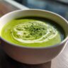 Close-up of a bowl of creamy asparagus soup, garnished with herbs and cream swirl.