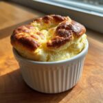 Close-up of a golden-brown cheese soufflé in a white ramekin, ready to eat. The image highlights the airy texture of the cheese soufflé.