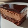 Close-up of a slice of Triple Chocolate Mousse Cake with a silky mirror glaze on a white plate.