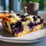 Close-up of a slice of Lemon Blueberry Cheesecake Dump Cake on a white plate, showing blueberries and cream.