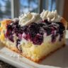 A slice of Lemon Blueberry Cheesecake Dump Cake on a white plate, topped with whipped cream.