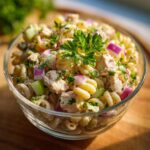 Close-up of a pasta salad and chicken in a glass bowl, with red onion, cucumber, and herbs.