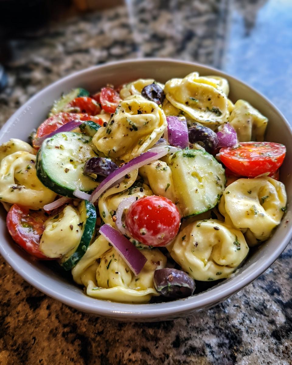 Close-up of a bowl of pasta salad tortellini with tomatoes, cucumbers, olives, and red onion.