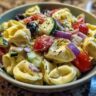 Close-up of a bowl of pasta salad tortellini with tomatoes, cucumbers, olives, and red onion.
