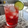 Close-up of The Driveway Drink, a refreshing red cocktail with ice and a lime wedge.
