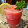 A glass of vibrant Vodka Strawberry Lemonade Slushy garnished with lemon and mint.